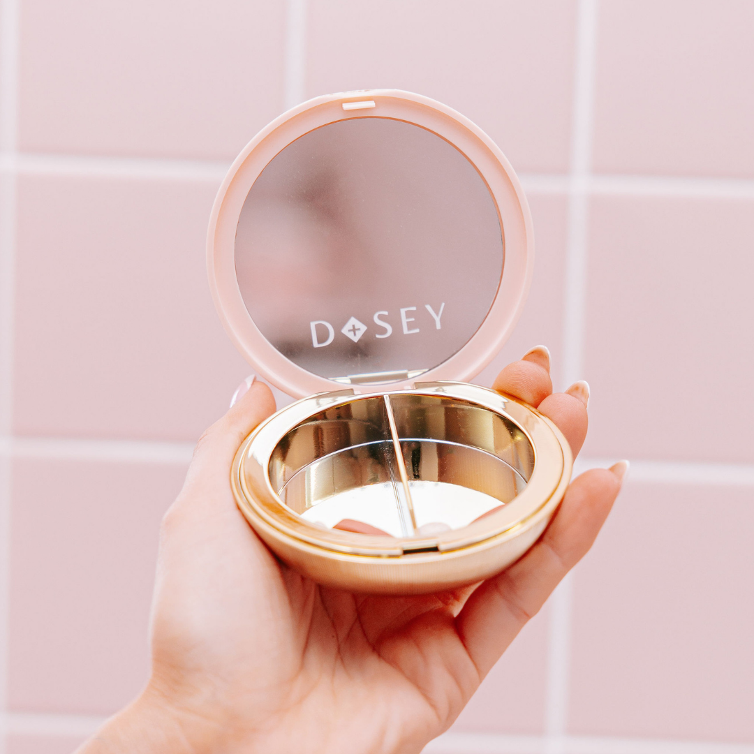 Hand holding a compact mirror with 'Dosey' branding against a pink tiled wall.