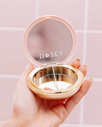 Hand holding a compact mirror with 'Dosey' branding against a pink tiled wall.