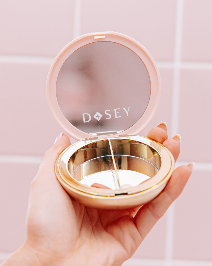 Hand holding a compact mirror with 'Dosey' branding against a pink tiled wall.